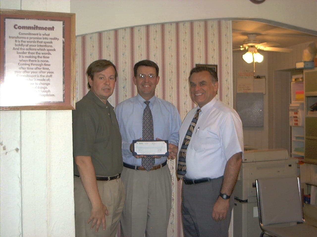 Three men hold up a check in an office, beside a framed poem titled Commitment