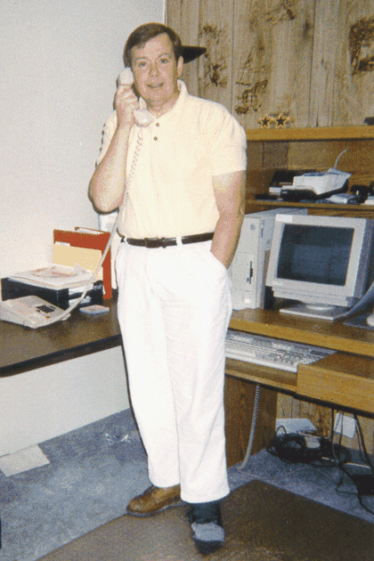 A man in a polo shirt talks on the phone beside an office desk with a computer