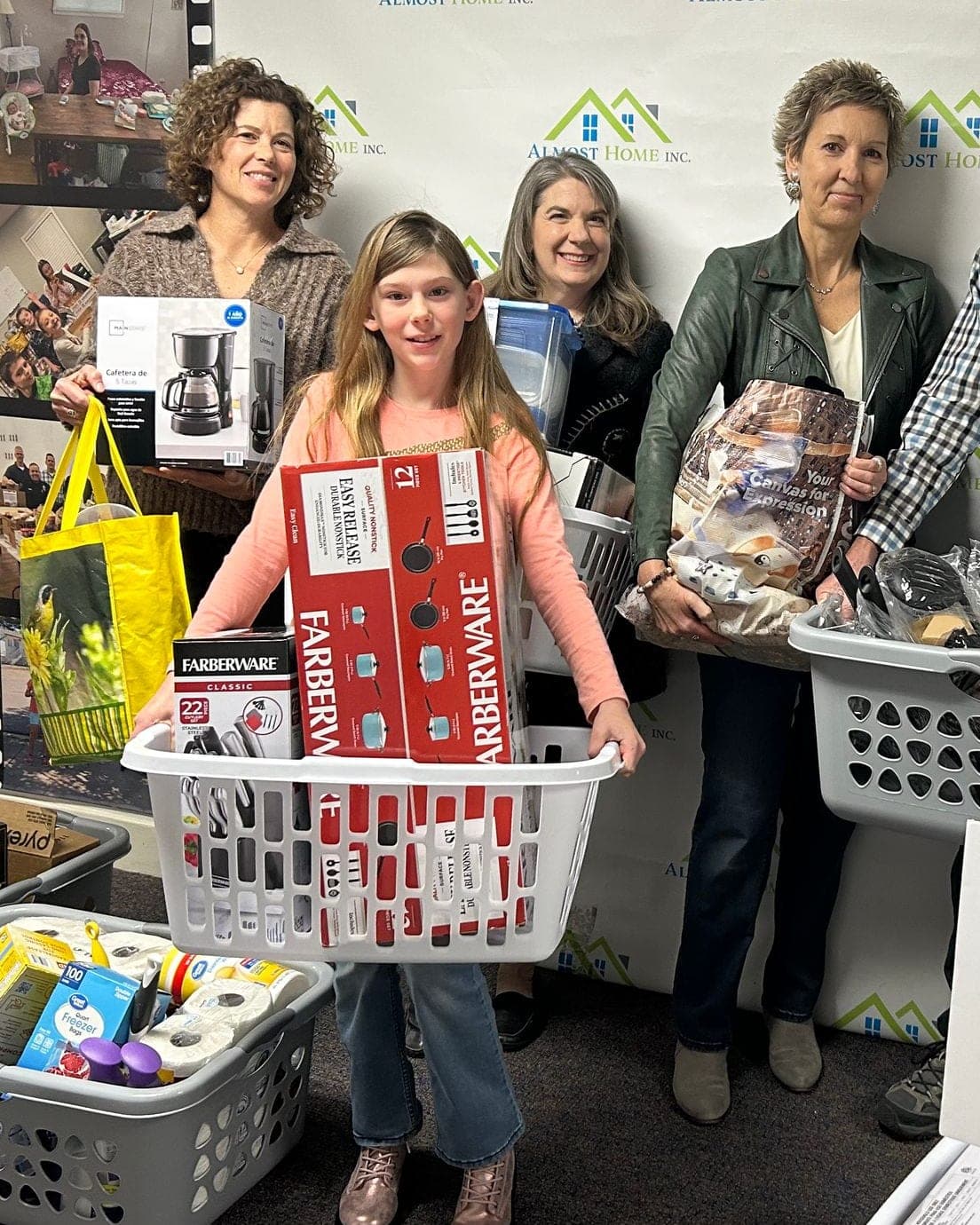 A girl and two women hold laundry baskets of cookware and household goods for Welcome Home baskets in front of the Almost Home logo wall