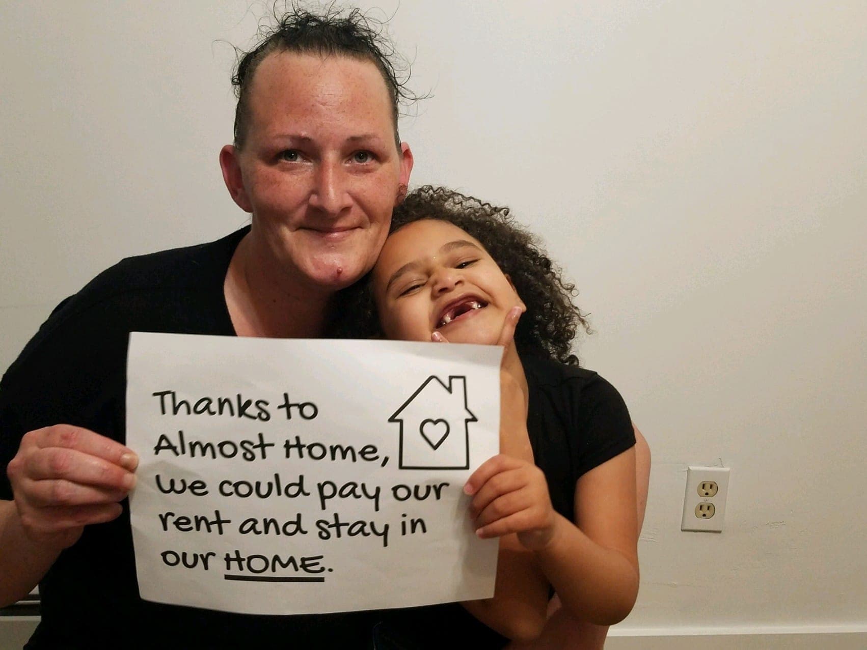 A mother and her laughing daughter hold a handwritten sign: Thanks to Almost Home, we could pay our rent and stay in our HOME.