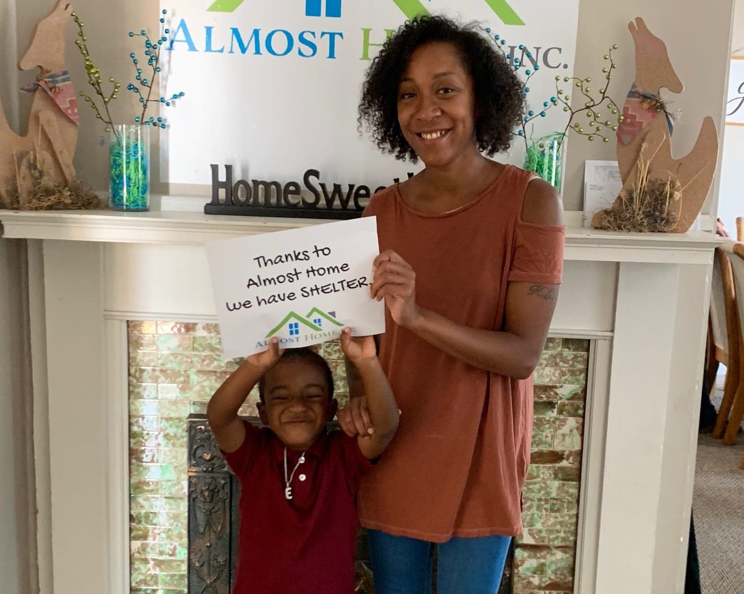 A mother and her young son by the family shelter mantel hold a sign: Thanks to Almost Home we have SHELTER.