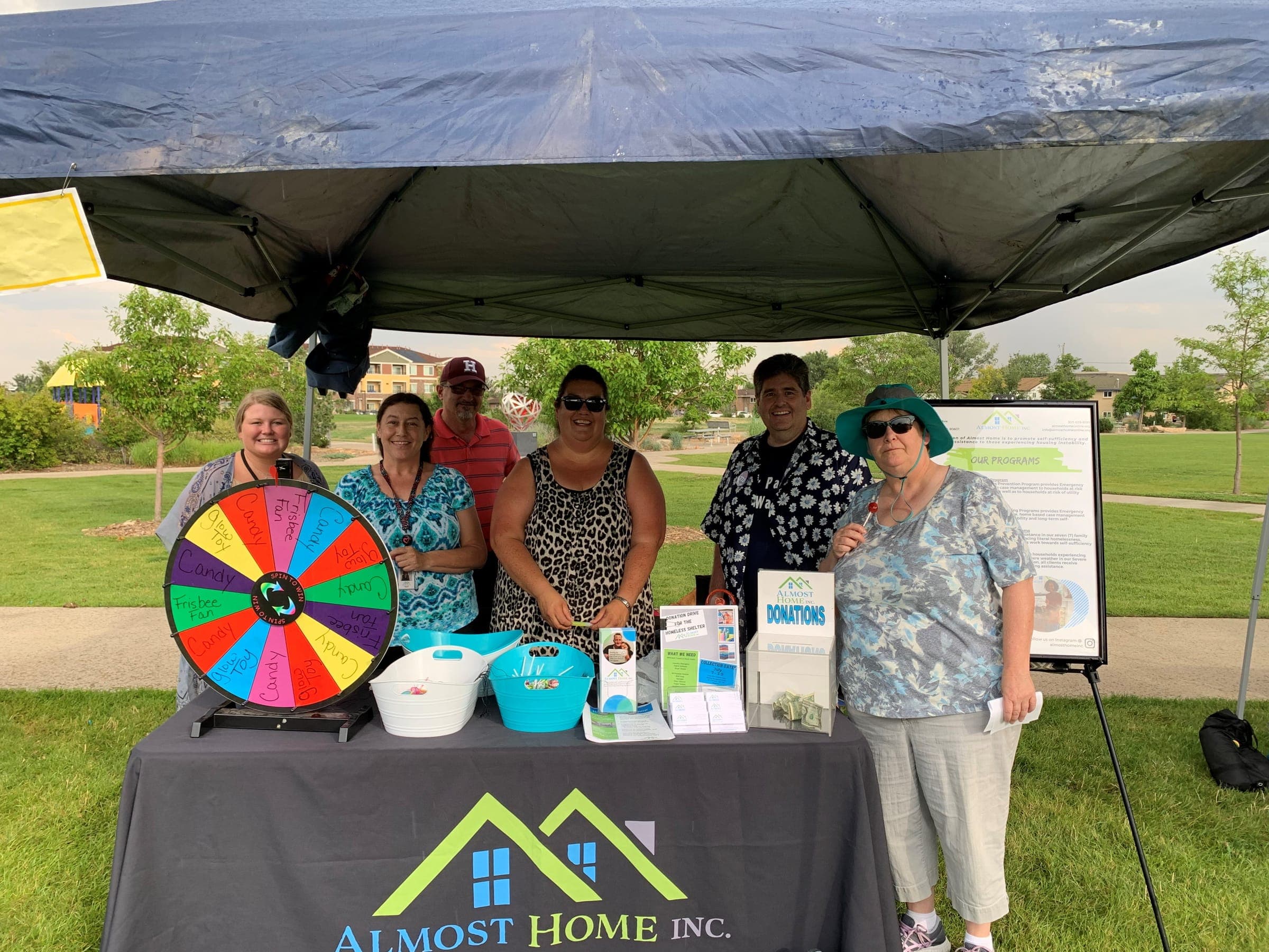 Six Almost Home staff and volunteers at an outdoor booth with the Almost Home tablecloth, a prize wheel and a donations box