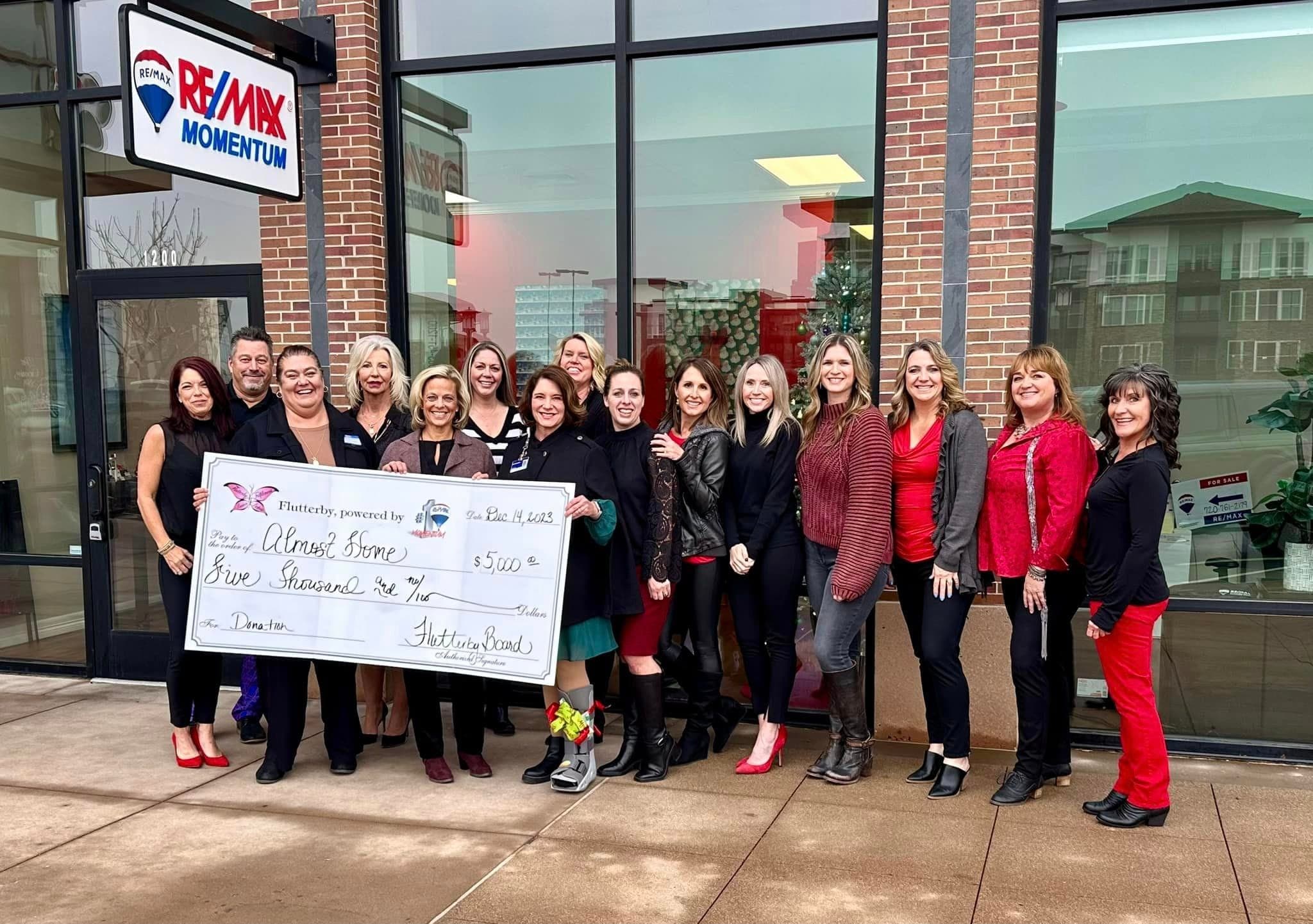 About seventeen people outside RE/MAX Momentum holding a $5,000 Flutterby check made out to Almost Home