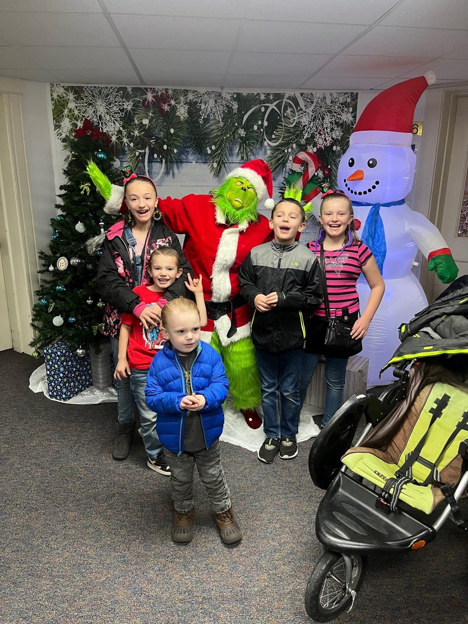 Five children pose with a Grinch at a family shelter holiday party, beside a Christmas tree
