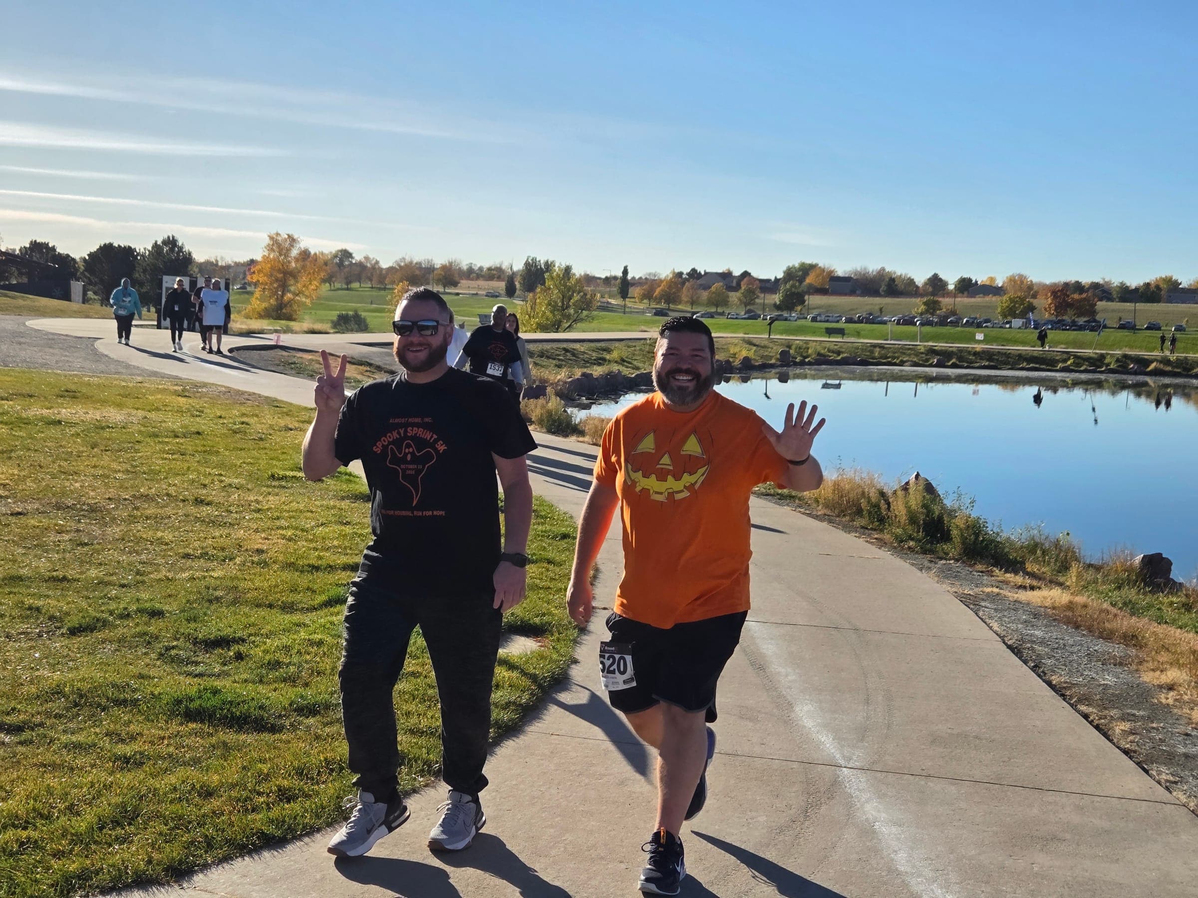 Two Spooky Sprint 5K runners smiling and waving on a lakeside path, one in the event T-shirt