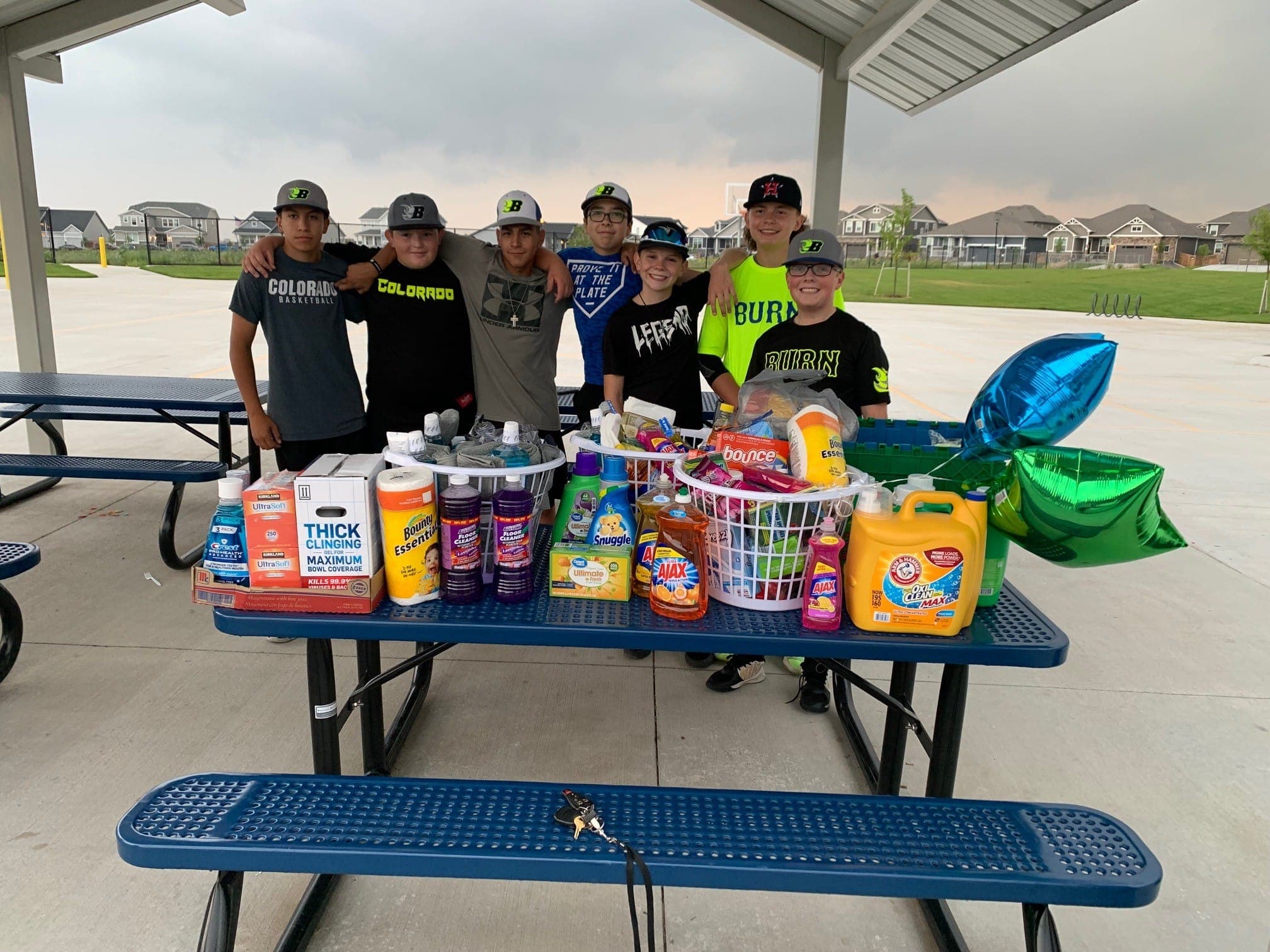 A youth baseball team behind a picnic table loaded with donated cleaning supplies and laundry baskets